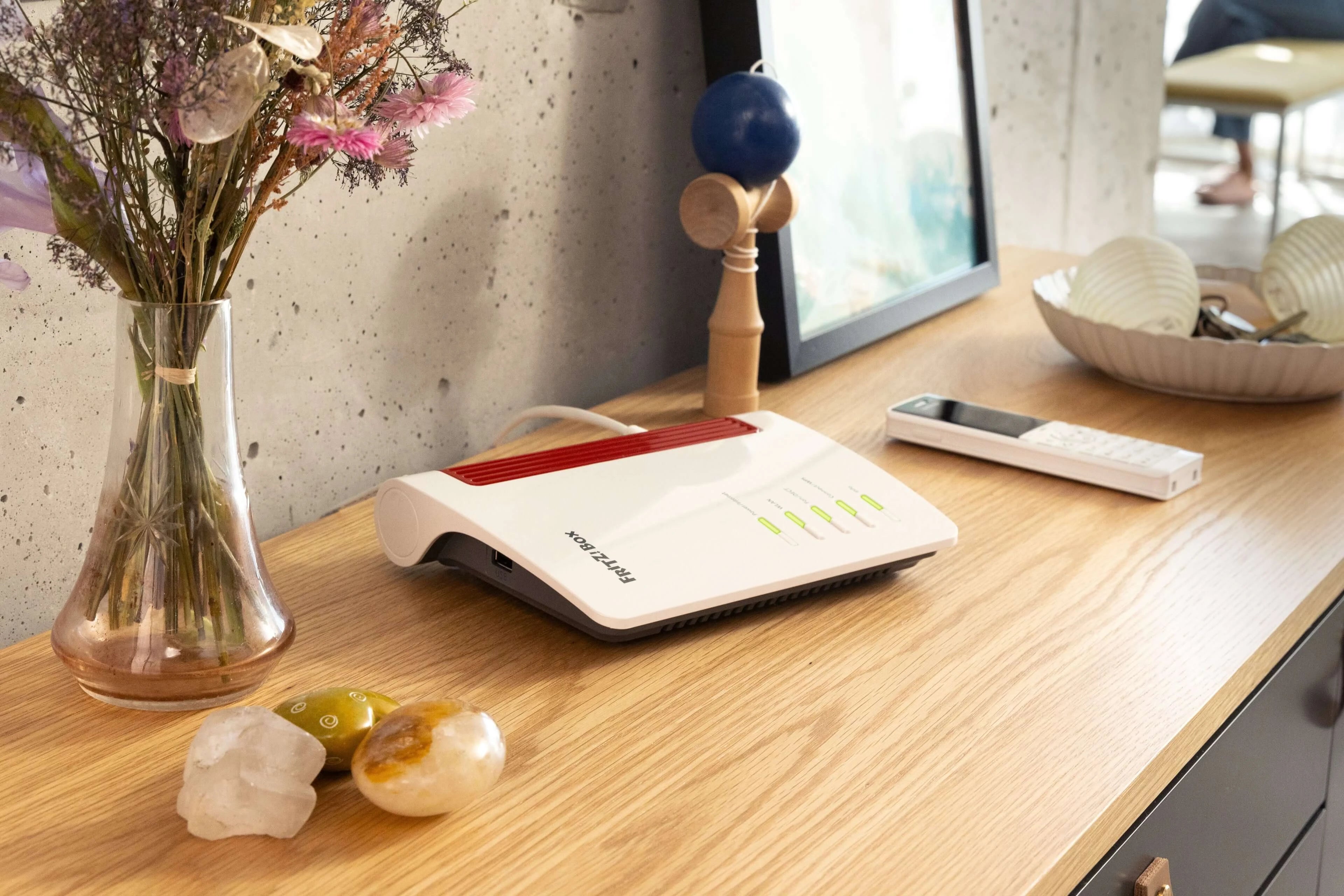 Ein moderner Router auf einem Holztisch mit Blumen, Steinen und einer Fernbedienung im Hintergrund.